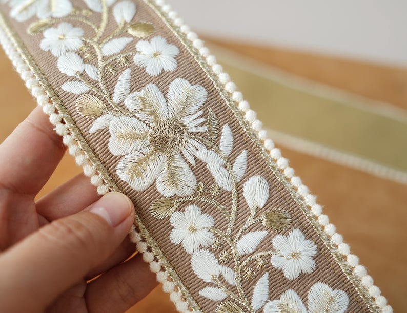 May include: Close-up of a decorative ribbon featuring embroidered white flowers and gold accents on a tan background. The ribbon is trimmed with a cream-colored pom-pom edge, suitable for various craft projects.