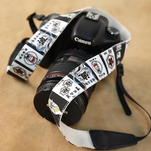 Könnte beinhalten: Eine schwarze Canon DSLR-Kamera mit einem Kameragurt, der einen weißen Hintergrund und ein wiederkehrendes Muster aus schwarz-weißen japanischen Grafiken aufweist, darunter stilisierte Figuren, Wellenmotive und verschiedene japanische Schriftzeichen.