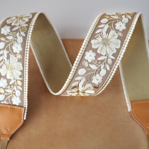 May include: A camera strap with a floral embroidered design. The strap features a tan background with white and gold floral embroidery. The edges are trimmed with a white beaded border. The ends have brown leather attachments.