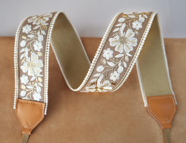 May include: A camera strap with a floral embroidered design. The strap features a tan background with white and gold floral embroidery. The edges are trimmed with a white beaded border. The ends have brown leather attachments.