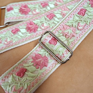 May include: Close-up of a guitar strap with a floral design. The strap features pink embroidered flowers and green leaves on a white background. The strap has a light brown leather end and a silver metal buckle.