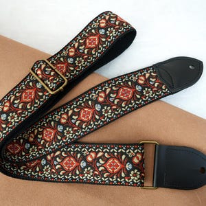 Op de afbeelding: Een gitaarband met een zwarte achtergrond en een herhalend bloemmotief in rood, wit en blauw. De band heeft zwarte leren uiteinden en messingkleurige metalen hardware voor aanpassing.