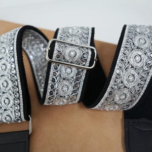 Könnte beinhalten: Ein schwarzer Gitarrengurt mit silbernen Stickereien. Der Gurt hat ein sich wiederholendes Blumenmuster mit einer silbernen Schnalle und schwarzen Lederenden. Entworfen für Gitarren oder andere Musikinstrumente.