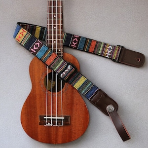 May include: A brown ukulele with a colourful patterned guitar strap. The strap has a brown leather end with a metal button.