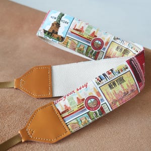 Könnte beinhalten: Ein weißer Canvas-Kameragurt mit einem braunen Leder-Endstück. Der Gurt ist mit einem bunten Druck von New Yorker Sehenswürdigkeiten und dem Text "New York" verziert.