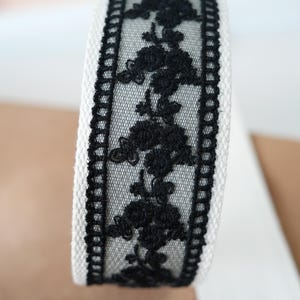 May include: Close-up of a roll of white fabric ribbon with a black floral lace overlay. The lace features a repeating pattern of stylized flowers. The edges of the ribbon are trimmed with a black border.