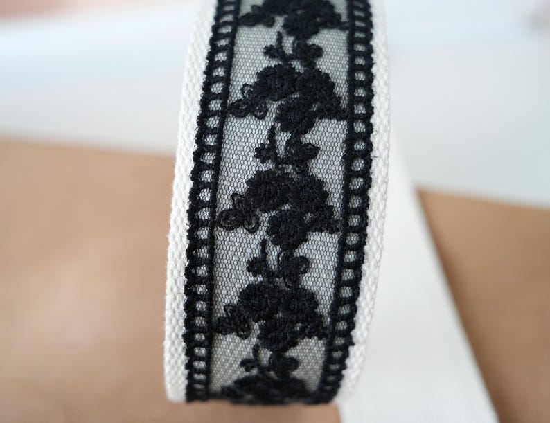 May include: Close-up of a roll of white fabric ribbon with a black floral lace overlay. The lace features a repeating pattern of stylized flowers. The edges of the ribbon are trimmed with a black border.