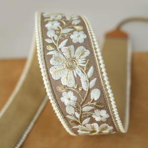 May include: A roll of decorative ribbon featuring a floral embroidered design. The ribbon has a tan base with a brown embroidered border. White and gold floral embroidery adorns the border, and a white beaded trim lines the edge.