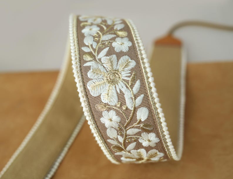 May include: A roll of decorative ribbon featuring a floral embroidered design. The ribbon has a tan base with a brown embroidered border. White and gold floral embroidery adorns the border, and a white beaded trim lines the edge.