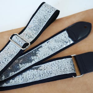 Könnte beinhalten: Ein Gitarrengurt mit schwarzem Rücken und silbernen Pailletten. Der Gurt hat schwarze Lederenden und eine silberne Metallschnalle zur Verstellung. Der Gurt wird auf einer braunen Oberfläche gezeigt.