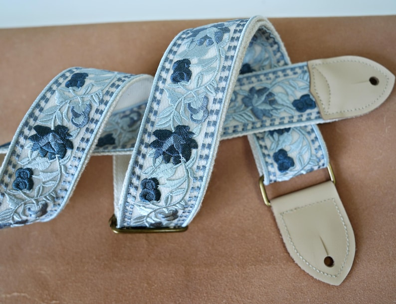以下が含まれることがあります： 水色と白のギター用ストラップ。濃い青色の花の刺繍模様が施されています。クリーム色の革製の端と真鍮色の金具が付いています。花の模様はストラップの長さに沿って繰り返されています。