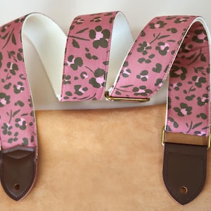 Könnte beinhalten: Ein rosa Gitarrengurt mit einem floralen Muster in Dunkelgrün und Hellrosa. Der Gurt hat braune Lederenden und goldfarbene Metallbeschläge. Die Innenseite des Gurtes ist weiß.