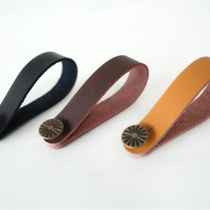 May include: Three leather drawer pulls in black, brown, and tan. Each pull features a loop design with a decorative, bronze-colored button. The pulls are shown against a white background, highlighting their colors and textures.