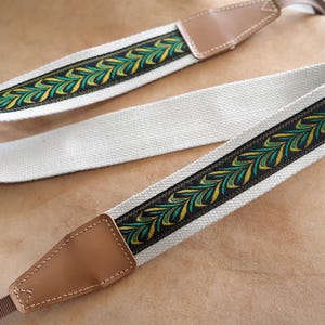 May include: A white canvas camera strap with a brown leather end piece and a green and gold patterned trim.