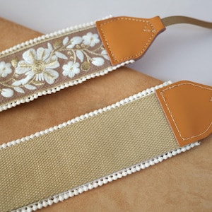 May include: Two camera straps. One strap features a tan fabric with white pom-pom trim and tan leather ends. The other strap has a brown fabric with white floral embroidery and white pom-pom trim, with tan leather ends.