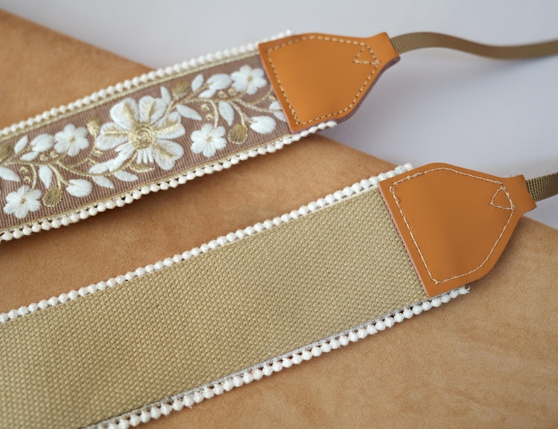 May include: Two camera straps. One strap features a tan fabric with white pom-pom trim and tan leather ends. The other strap has a brown fabric with white floral embroidery and white pom-pom trim, with tan leather ends.