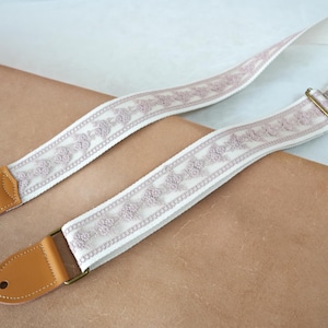 May include: A cream guitar strap featuring a mauve floral design. The strap has brown leather ends and brass-coloured metal hardware. The design is elegant and suitable for acoustic or electric guitars. The strap's length is approximately 150 cm.