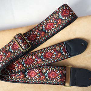 Könnte beinhalten: Ein Gitarrengurt mit einem detaillierten, farbenfrohen Muster. Der Gurt hat einen schwarzen Hintergrund mit einem sich wiederholenden Design in Rot, Orange, Blau und Weiß. Er hat schwarze Lederenden und messingfarbene Hardware.
