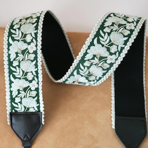 May include: A camera strap with a dark green floral embroidered design. The strap features white flowers and leaves with a white scalloped edge. The strap has black leather ends and a black interior.
