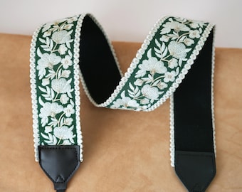 May include: A camera strap with a dark green floral embroidered design. The strap features white flowers and leaves with a white scalloped edge. The strap has black leather ends and a black interior.