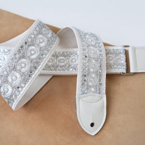 May include: A white guitar strap featuring an intricate silver embroidered pattern with circular motifs and small sequins. The strap includes white leather end tabs with pre-cut holes for attachment and silver-toned metal hardware adjusters.