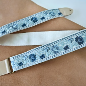 以下が含まれることがあります： クリーム色の中心部と水色の花の刺繍が施されたギターストラップ。濃い青色の花のアクセントと、真鍮色の金具が付いた薄茶色の革製の端が特徴です。