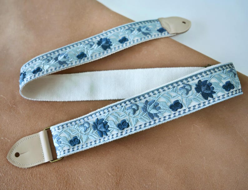 以下が含まれることがあります： クリーム色の中心部と水色の花の刺繍が施されたギターストラップ。濃い青色の花のアクセントと、真鍮色の金具が付いた薄茶色の革製の端が特徴です。