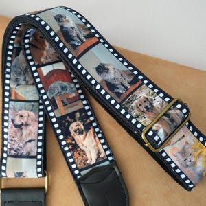 May include: Black guitar strap featuring a repeating pattern of pet photos, including cats and dogs. The strap has a black leather end and gold-colored metal hardware. The strap is approximately 2 inches wide.