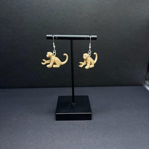 May include: A pair of dangle earrings featuring tan monkey figures. The earrings are suspended from silver-colored hooks. The monkeys are in a seated position with their arms outstretched. The background is a solid black.
