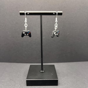 May include: A pair of silver dangle earrings with black game controller charms. The charms have silver buttons and are hanging from silver hooks.