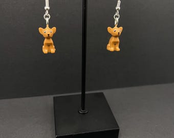 May include: A pair of dangle earrings featuring a brown Lego chihuahua figurine on each earring. The earrings are hanging from a black earring stand.