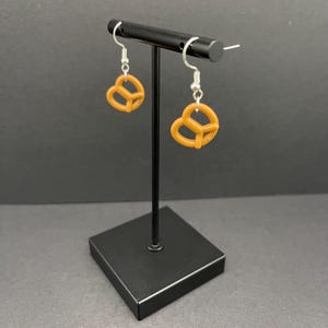May include: A pair of dangle earrings featuring a brown pretzel shape. The earrings are hanging from a black earring stand.