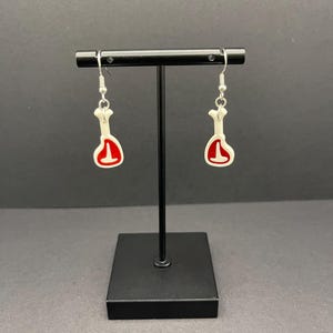 May include: A pair of silver dangle earrings with white and red bone-shaped charms. The charms are shaped like a cut of meat.