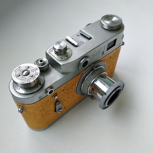 May include: A vintage FED 3 rangefinder camera featuring a silver metal top plate and a textured mustard yellow body. The camera includes a silver lens with knurled focus rings, various adjustment dials with numerical markings, and a shutter button.
