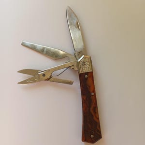 May include: A multi-tool pocket knife featuring a brown marbled handle with a metallic bolster engraved with a leaf pattern. The tool includes a main blade, a pair of scissors, and a flat-head screwdriver or bottle opener attachment.