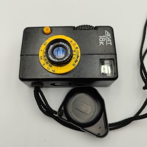 May include: A black Agat 18K film camera featuring a bright yellow lens ring with aperture and weather icons, a yellow shutter button, and a black lens cap attached by a black cord. The camera body displays the text Agat 18K in white.