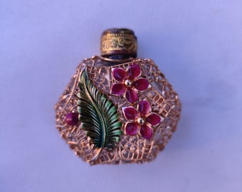 Op de afbeelding: Een klein, decoratief parfumflesje met een goudkleurige, ingewikkelde filigraan metalen kooi. De buitenkant is versierd met een groen geëmailleerd blad en twee felroze geëmailleerde bloemen. Het flesje heeft een cilindervormige, textuurrijke gouden dop.