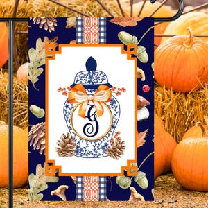 May include: A blue and white porcelain ginger jar with a gold monogram "G" and an orange bow. The jar is surrounded by fall-themed elements like acorns, pine cones, and leaves. The background is a dark blue with a white and orange plaid pattern.