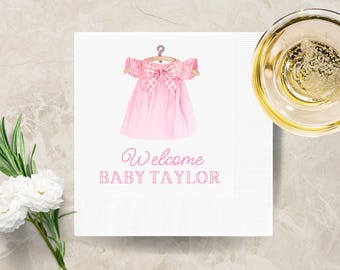 May include: White baby shower napkin with a watercolor illustration of a pink dress on a hanger. The text "Welcome BABY TAYLOR" is printed in pink script. A glass of champagne and flowers are in the background.