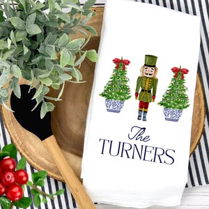 May include: White kitchen towel with a watercolor illustration of a nutcracker and two Christmas trees decorated with lights and ornaments. The text "The TURNERS" is printed in blue below the illustration.