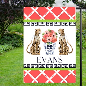 May include: A white garden flag with a red and white lattice pattern, a green and blue border, and a blue and white ginger jar with pink flowers. Two cheetah silhouettes are on either side of the ginger jar. The text "EVANS" is printed in black below the ginger jar.