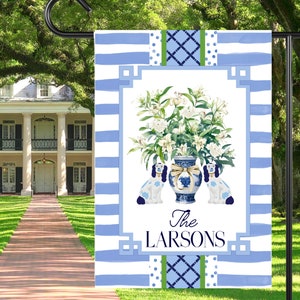 May include: A blue and white garden flag with a floral design and two dogs flanking a vase. The text "The Larsons" is at the bottom of the flag.