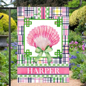 May include: A garden flag featuring a pink seashell design with a green bow and the name "HARPER" on a pink banner. The flag has a plaid pattern in pink, green, and navy blue. The flag is displayed in a garden setting.