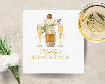 May include: White paper napkin with a champagne bottle and two glasses illustration. The bottle is adorned with flowers and tied with a gold ribbon. The text "Maddy's BRIDAL SHOWER" is in gold script. A glass of champagne is in the upper right corner.