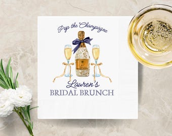 May include: White paper napkin with a watercolor illustration of a champagne bottle and two flutes. The text "Pop the Champagne" and "Lauren's Bridal Brunch" are printed in navy blue.