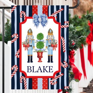 May include: A blue and red striped Christmas garden flag with a white panel featuring two nutcracker figurines, a potted evergreen tree, and the name "BLAKE".