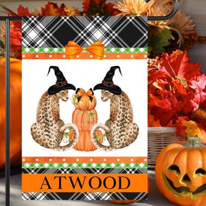 May include: A rectangular garden flag featuring a black and white plaid border, an orange bow, and two leopards wearing black witch hats flanking a stack of three orange pumpkins. The bottom orange band displays the black text ATWOOD.