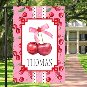 May include: A pink and white garden flag with a red border. The flag features a white background with two red cherries with a pink bow on top. The cherries are on a white background with a red border. The text "THOMAS" is printed in black below the cherries. The flag is surrounded by a pink background with a pattern of red cherries.