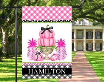 May include: Pink and green garden flag featuring a stack of pink pumpkins in a white urn, flanked by two pink pineapples. The flag has a black and white checkered border with the name "HAMILTON" in black text.