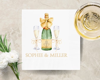 May include: White paper napkin with a watercolor illustration of a champagne bottle and two glasses. The bottle is green with a gold bow and a fleur-de-lis. The text "SOPHIE & MILLER" is in gold.